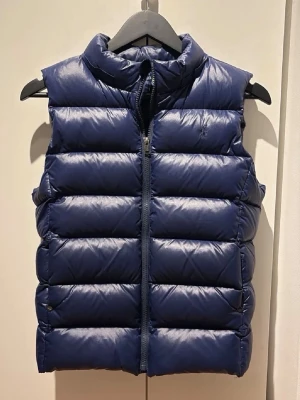 Mörkblå dunväst med hög krage - Snygg mörkblå dunväst med hög krage och quiltad design. Västen har dragkedja framtill och breda kanaler som ger en puffig look. Perfekt att slänga över en hoodie eller tröja för extra värme. Enkel och stilren modell utan ärmar.