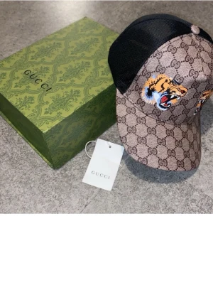 Tiger Gucci keps  - Snygg Gucci keps med klassiskt GG-mönster i beige och brunt, tigerbrodyr framtill och svart mesh baktill. Justerbar rem med grönt och rött band. Märkesdetaljer på insidan och etikett medföljer. Perfekt för dig som vill sticka ut. 