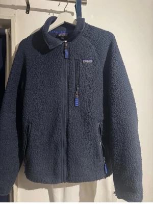 Mörkblå teddyjacka från Patagonia - Mysig mörkblå teddyjacka från Patagonia med hög krage och tre dragkedjeförsedda fickor. Jackan har en fluffig, fårskinnsliknande yta och detaljer i blått vid dragkedjorna. Perfekt för kalla dagar när du vill vara både varm och stilren.