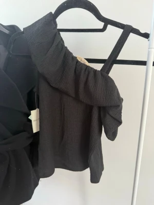 Svart asymmetrisk topp med volang - Trendig svart topp med en axel och bred volangdetalj längs halsringningen. Tillverkad i ett strukturerat, lätt stretchigt tyg som ger en cool och modern look. Perfekt för dig som vill sticka ut med en unik och stilren topp.