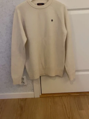 Beige stickad tröja från Morris - Klassisk beige stickad tröja från Morris. med rund halsringning och diskret logga broderad på bröstet. Tröjan har ribbstickade muddar vid ärmslut och nederkant, perfekt för lager på lager och en clean look.