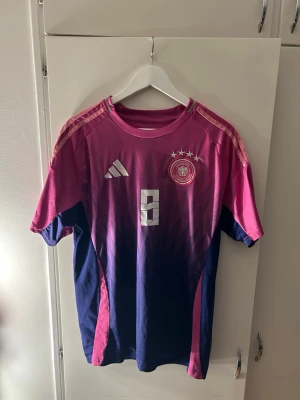 Tysklands matchtröja Kroos #8 Adidas - Snygg Tyskland matchtröja från Adidas i rosa och mörkblått med nummer 8 och Kroos på ryggen. Tröjan har tre stjärnor ovanför landslagsemblemet, klassiska Adidas-ränder på axlarna och är tillverkad i lätt, ventilerande material. Perfekt för fotbollsträning eller som supporterplagg samt att ha på fritiden!