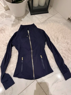 Marinblå jacka från Lululemon - Snygg marinblå jacka från Lululemon med figurnära passform och guldfärgade dragkedjor. Jackan har hög krage, långa ärmar och två fickor framtill. Tillverkad i ett stretchigt funktionsmaterial som passar perfekt till vardags eller träning.