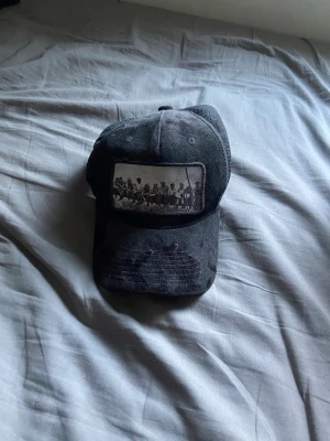 Svart keps med vintage tryck - Säljer en svart truckerkeps med mesh på sidorna och böjd skärm. Framtill finns ett vintage-inspirerat tryck med svartvit bild på ett gäng personer. Kepsen har justerbar snapback och är tillverkad i bomull och mesh. Perfekt för dig som gillar retrostil.