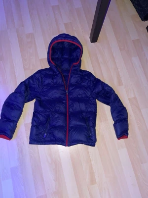 Blå dunjacka med röd dragkedja Polo - Snygg blå dunjacka från Polo Ralph Lauren med röd dragkedja och röda detaljer vid huva och ärmslut. Jackan har två fickor med dragkedja och broderad logga på bröstet. Perfekt för kalla vinterdagar och riktigt skön att bära.