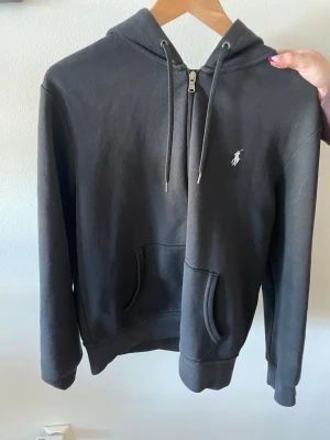 Svart hoodie från Polo Ralph Lauren - Klassisk svart hoodie med dragkedja från Polo Ralph Lauren. Har huva med dragsko, känguruficka och den ikoniska vita logotypen broderad på bröstet. Tillverkad i mjukt bomullsmaterial som är skönt att bära.