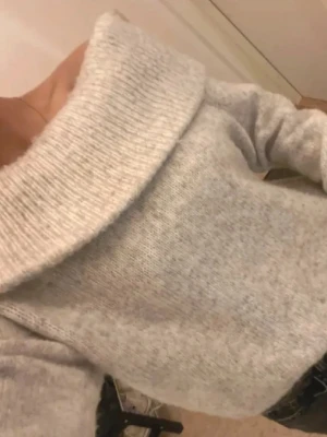 Ljusgrå off shoulder stickad tröja - Mysig ljusgrå stickad tröja med bred off shoulder-krage. Tröjan har en avslappnad passform och långa ärmar, perfekt för lager på lager. Materialet ser ut att vara mjukt och skönt, troligen en ull- eller akrylblandning. Passar sofrlek S med 