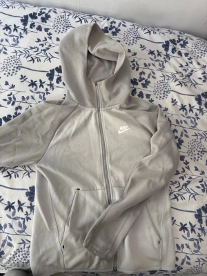 Ljusgrå zip hoodie från Nike (Sällsynt) - Snygg ljusgrå hoodie från Nike med dragkedja framtill och två fickor. Klassisk Nike-logga på bröstet och huva för extra stilpoäng. Tillverkad i mjukt material som känns skönt mot huden. Perfekt för dig som gillar sportig och avslappnad look.