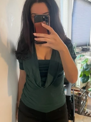 Mörkgrön topp med draperad urringning - Snygg mörkgrön topp med korta ärmar och draperad v-ringad urringning. Toppens material känns mjukt och stretchigt, perfekt för en trendig look. Passformen är normal och toppen är enkel att matcha med olika outfits.