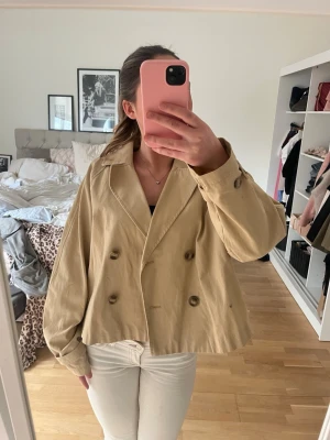 Kort beige trenchcoat - Snygg kort trenchcoat i beige med klassisk dubbelknäppning och bred krage. Den har små tecken på användning då den har ett litet smuts märke på ärmen och framsidan!💞storlek L men passar som en M tycker jag! kontakta vid frågor eller köp direkt😊