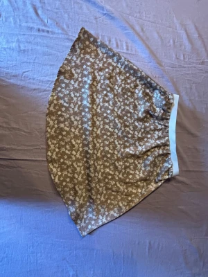 Brun blommig kortkjol med resår - Söt brun kortkjol med vitt blommönster och bred resår i midjan. Kjolen har en lätt utsvängd form och är perfekt för dig som gillar en avslappnad men ändå söt stil. Materialet känns mjukt och lätt, vilket gör den skön att bära under varmare dagar.