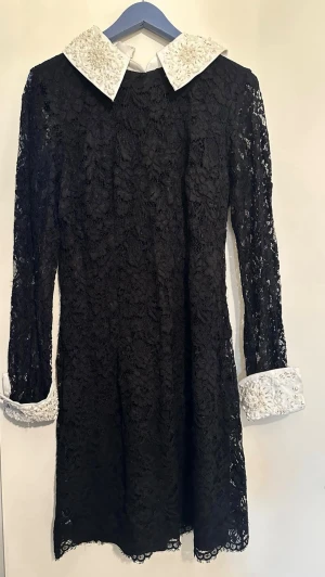 Svart spetsklänning med pärlkrage - Elegant svart spetsklänning med lång ärm och vit krage samt manschetter dekorerade med pärlor. Klänningen har dragkedja i ryggen och är knälång. Perfekt för dig som vill ha en chic och unik look.ena ärmen har tappat några stenar men påverkar inte funktionen. Liten i storleken, mer som M/L än XL 