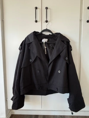 Kort svart trenchcoat  - Snygg svart trenchcoat i kort modell med dubbelknäppning och bred krage. Från Josefine HJs kollektion med NA-KD. Den perfekta vårjackan!! 