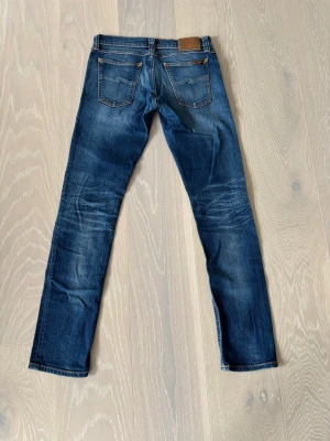 Nudie Jeans  - Säljer ett par klassiska blå jeans från Nudie Jeans med smal passform och snygga slitningar. Jeansen har fem fickor, kontrastsömmar och en läderpatch bak i midjan. Tillverkade i denim med en cool tvättad look. Vid intresse är det bara att höra av sig. Pris går att diskutera 