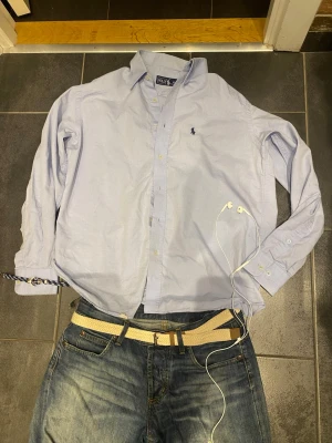 Ljusblå skjorta från Polo Ralph Lauren - Klassisk ljusblå skjorta från Polo Ralph Lauren med button-down krage och broderad logga på bröstet. Skjortan har lång ärm, knappar framtill och är tillverkad i bomull. Perfekt för en clean och stilren look. Skulle säga att den är M/L (passar också dem som har L). 