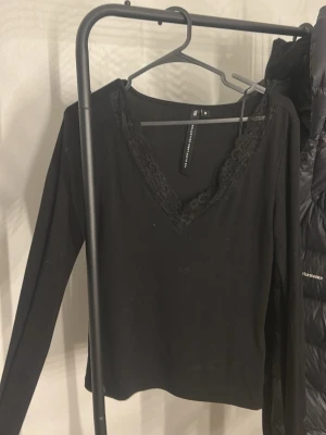 Svart långärmad topp med spets - Svart långärmad topp från The Black Label Collection med v-ringning och fina spetsdetaljer runt halsen. Perfekt att styla med jeans eller kjol för en snygg look. Mjuk och stretchig känsla.