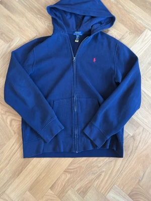 Marinblå hoodie från Polo Ralph Lauren - Snygg marinblå hoodie med dragkedja från Polo Ralph Lauren. Klassisk design med huva, kängurufickor och den ikoniska röda loggan broderad på bröstet. Tillverkad i mjuk bomull för skön komfort och avslappnad stil. Är för åldern 18-20 vilket ungefär motsvarar S i herr.