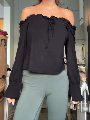 Svart offshoulder topp med långa ärmar - Trendig svart offshoulder topp med långa ärmar och smockad överdel. Toppen har en söt knytning framtill vid halsen och är croppad i modellen. Perfekt för dig som gillar en stilren och modern look.