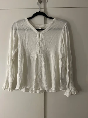 Vit långärmad blus med knappar - Säljer en vit långärmad blus med v-ringning och små knappar framtill. Blusen har ett lätt crinklat tyg och volang vid ärmslut och nederkant. Perfekt för en avslappnad men ändå stilren look. Storlek.