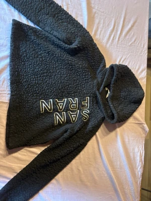 Svart teddyjacka med huva SAN FRAN - Mysig svart teddyjacka med huva och stora vita bokstäver 'SAN FRAN' på framsidan. Jackan har en fluffig, fårullsliknande struktur och långa ärmar. Perfekt för kalla dagar när du vill vara både varm och trendig.