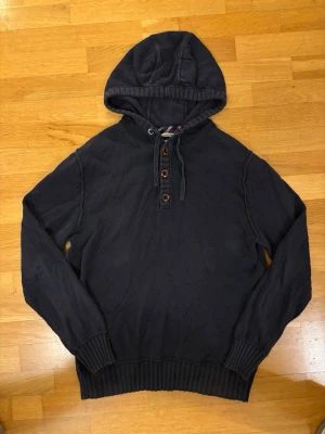 Mörkblå hoodie med knappar - Säljer en mörkblå hoodie med huva och fyra träknappar framtill. Hoodien har ribbade muddar vid ärmslut och nederkant samt en avslappnad passform. Materialet är mjukt och ser ut att vara bomull eller bomullsblandning. Perfekt för en chill och stilren look.