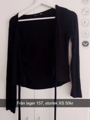 Svart omlottkofta från Lager 157 - Svart omlottkofta från Lager 157 med långa ärmar och knytband i midjan. Enkel och stilren design som passar perfekt till olika outfits. Tillverkad i mjukt och stretchigt material för en bekväm känsla.