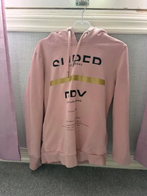 Rosa hoodie med tryck och huva - Snygg rosa hoodie från Superdry med svart och guldigt tryck framtill. Hoodien har en klassisk huva med dragsko och ribbade muddar vid ärmar och nederkant. Perfekt för en avslappnad och trendig look.