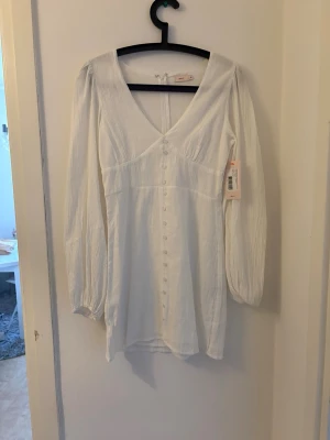 Vit kortklänning med knappar - Snygg vit kortklänning med v-ringning och långa ärmar. Klänningen har dekorativa knappar framtill och lätt puffade axlar. Tillverkad i ett tunt, strukturerat tyg som ger en luftig känsla. Perfekt för sommarens alla tillfällen.