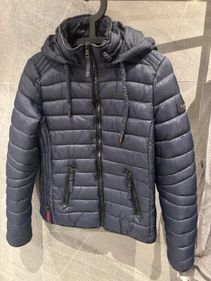 Mörkblå pufferjacka från Navahoo - Snygg mörkblå pufferjacka från Navahoo med huva och dragsko, två dragkedjeförsedda fickor och quiltad design. Jackan har en sportig look och är perfekt för kalla vinterdagar. Materialet är glansigt och ger en modern vibe.