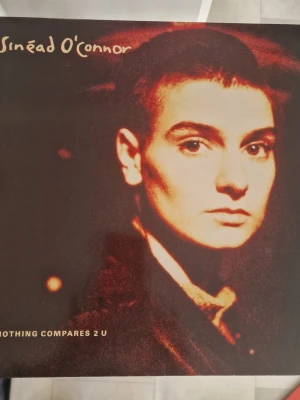 Sinéad O'Connor vinyl Nothing Compares 2 U - Vinylskiva med Sinéad O'Connor och låten 'Nothing Compares 2 U'. Omslaget är mörkrött och svart med artistens namn och låttitel i gul text. Baksidan är vinröd med låtlista och skivbolagets logga. Perfekt för samlare av klassisk musik på vinyl. Inga repor.