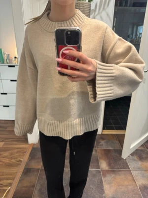 Beige oversized stickad tröja - Säljer en beige oversized stickad tröja med breda ärmar och ribbade muddar vid hals, ärmslut och nederkant. Tröjan har en avslappnad passform och är perfekt för lager på lager. Materialet känns mjukt och mysigt, men exakt material är okänt.