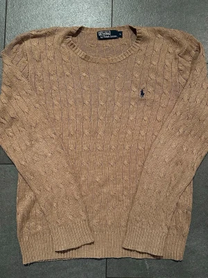Beige kabelstickad tröja Polo Ralph Lauren - Kabelstickad beige tröja från Polo Ralph Lauren med rund halsringning och klassisk broderad logga på bröstet. Tröjan har långa ärmar och ribbade muddar vid ärmslut och nederkant. Perfekt för lager på lager och snygg till jeans.