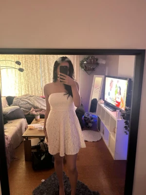 Vit axelbandslös spetsklänning - Säljer en söt vit axelbandslös klänning med spetsdetaljer och utställd kjol. Klänningen är kort och har en feminin, romantisk look som passar perfekt till sommarens alla tillfällen. Materialet känns lätt och luftigt. Perfekt studentklänning storlek 38 men passar mig som vanligtvis är storlek 36