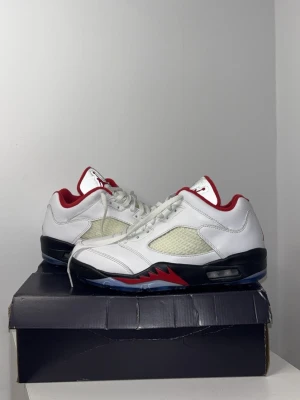 Air Jordan 5 Low Fire Red Golf sneakers - Säljer ett par Air Jordan 5 Low Fire Red sneakers i vitt läder med svarta och röda detaljer. Skorna har genomskinlig sula, meshpaneler på sidorna och röd Jumpman-logga på plösen. Röd insida och svarta sulor med röda accenter ger en clean och sportig look.