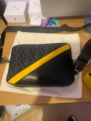 Fendi axelväska svart/gul - Snygg axelväska från Fendi i svart läder med en diagonal gul detalj och klassiskt FF-mönster på ovansidan. Väskan har bred justerbar axelrem med Fendi-logga och silverfärgade metalldetaljer. Perfekt för dig som vill ha en trendig och exklusiv accessoar.