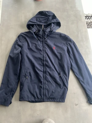 Marinblå vindjacka från Polo Ralph Lauren - Snygg marinblå vindjacka från Polo Ralph Lauren med huva och dragkedja framtill. Invik bar luva och jackan är kemtvättad. Skick 9/10, priset kan diskuteras. Jackan har två snedställda fickor med dragkedja och den klassiska röda logotypen broderad på bröstet. Jackan är en tjockare typ av vindjacka 