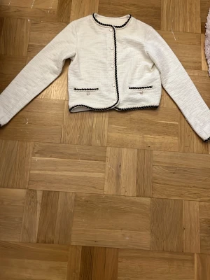 Vit cardiganjacka med svarta detaljer - Stilren vit cardiganjacka med svarta kantband och dekorativa fickor framtill. Jackan har rund halsringning och är tillverkad i ett strukturerat tyg som ger en exklusiv känsla. OBS det är barn storlek men skulle säga att det är ungefär xxs-xs 