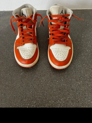 Nike Air Jordan 1 Mid orange/vit - Säljer ett par Nike Air Jordan 1 Mid sneakers i orange och vitt med svarta detaljer. Skorna har klassisk swoosh på sidan, perforerad tåbox och högt skaft. Tillverkade i läder. Skorna är i storlek 39 och är i bra skick!