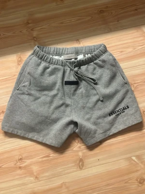 Grå shorts från Essentials - Säljer ett par grå mjukisshorts från Essentials med dragsko i midjan och två sidofickor. Shortsen har en relaxed fit och logga på vänster ben samt en svart patch framtill. Perfekta för chill dagar och streetwear-stil.