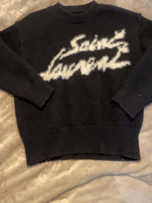 Svart stickad Saint Laurent-tröja - Cool svart stickad tröja från Saint Laurent med vit, grafisk logga framtill. Tröjan har rund halsringning, långa ärmar och ribbade muddar vid ärmslut och nederkant. Perfekt för dig som gillar streetstyle och designerplagg.