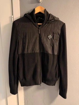 Svart quiltad jacka från J.Lindeberg - Snygg svart jacka från J.Lindeberg med quiltad framsida och huva. Jackan har dragkedja framtill, ribbade muddar och en diskret logga på bröstet. Materialmix med stickade ärmar och nederdel samt polyester på överdelen. Perfekt för  vår dagar. Pris kan diskuteras 