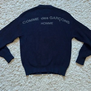 CGD | Knit Sweater  - Säljer en mörkblå stickad tröja från Comme des Garçons Homme i storlek S. Tröjan har ett grovt ribbstickat material, rund hals och tryckt logga i grått på ryggen. Perfekt för dig som gillar clean design och streetwear-vibe. Den är väldigt varm och cozy att ha på sig. Passar helt perfekt runt axlarna och armarna- ett av mina favorit plagg som jag säljer! 