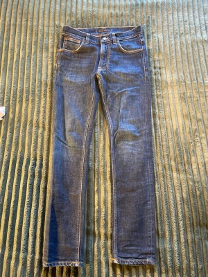 Blå jeans från Nudie Jeans - Säljer ett par klassiska blå jeans från Nudie Jeans med fem fickor, kontrastsömmar och rå kant vid benslut. Jeansen har en smal passform och är tillverkade i fint material. Perfekta till en avslappnad stil. W29 L30