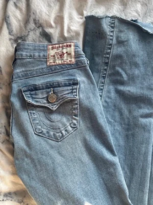 Ljusblå True Religion jeans - Säljer ett par ljusblå jeans från True Religion med ikoniska ficklock och silverfärgade knappar bak. Jeansen har raka ben och klassisk femficksdesign. Snygga kontrastsömmar och patch med logga bak i midjan. Perfekta för en avslappnad streetstil.