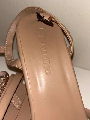 Beige sandaler med glitterdetalj - Säljer ett par stilrena beige sandaler från Excellence med fyrkantig tå och smala remmar, varav en med glitterdetalj. Klackarna är blockformade och remmarna är i syntetmaterial. Perfekta för dig som gillar en clean look med lite extra bling.