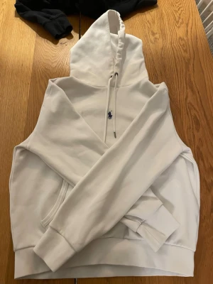 Vit hoodie från Polo Ralph Lauren - Säljer en clean vit hoodie från Polo Ralph Lauren med klassisk huva och dragsnören. Den har en broderad marinblå logga på bröstet och en stor magficka framtill. Hoodien är i mjukt bomullsmaterial och har ribbade muddar vid ärmar och nederkant.