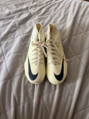  Nike fotbollsskor med dobbar - Säljer ett par  fotbollsskor från Nike med högt skaft och svarta detaljer, inklusive den klassiska swoosh-loggan och stor NIKE-text på sidan. Skorna har snörning och fasta dobbar för bra grepp på planen. Perfekta för dig som vill sticka ut på matchen.