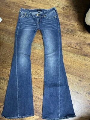 Mörkblå true religion low - Snygga blå true religion bootcut jeans med lätt slitna detaljer och klassisk femficksdesign. Jeansen har en låg midja och utsvängda ben, vilket ger en retro vibe. Tillverkade i denim med kontrastsömmar och dragkedjegylf.