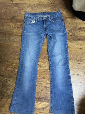 Blå bootcut jeans med slitningar - Ett par klassiska blå bootcut jeans med lätt slitna detaljer framtill och bakfickor. Jeansen har normal low  midja, fem fickor och stängs med dragkedja och knapp. Perfekta till sneakers eller boots för en avslappnad look.