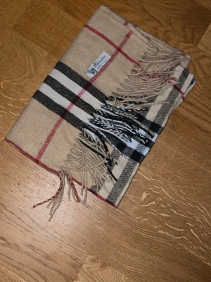 Beige rutig halsduk från Burberry - Klassisk beige halsduk från Burberry med ikoniskt rutmönster i svart, vitt och rött. Halsduken har fransar i ändarna och är tillverkad i mjukt material som känns lyxigt. Perfekt accessoar för att lyfta din outfit.